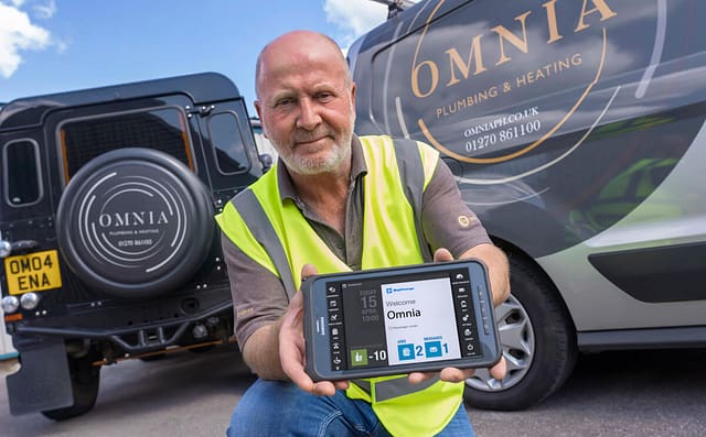 Omnia plumber on front of van holding up a BigChange tablet to camera.